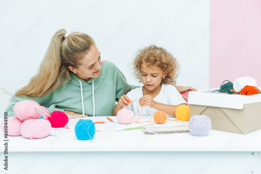 Mom teaches her daughter to knit. Mom and a little blonde girl are sitting at the table and crocheting. Mom teaches her daughter to knit. Mom and a little blonde girl are sitting at the table and crocheting.