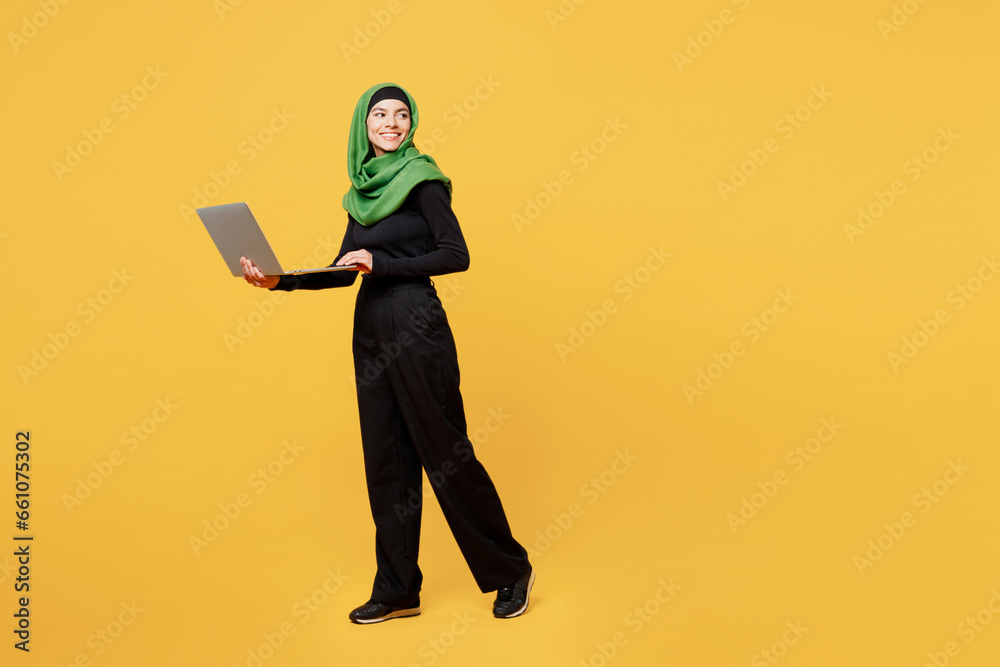 Full body young arabian asian muslim IT woman wears green hijab abaya black clothes work hold use laptop pc computer look aside isolated on plain yellow background. People uae islam religious concept. Full body young arabian asian muslim IT woman wears green hijab abaya black clothes work hold use laptop pc computer look aside isolated on plain yellow background. People uae islam religious concept.