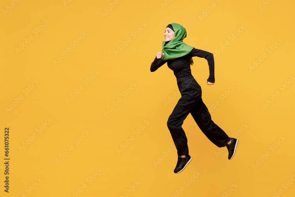 Full body side profile view young arabian asian muslim woman wear green hijab abaya black clothes jump high run fast hurry up isolated on plain yellow background. People uae islam religious concept. Full body side profile view young arabian asian muslim woman wear green hijab abaya black clothes jump high run fast hurry up isolated on plain yellow background. People uae islam religious concept.