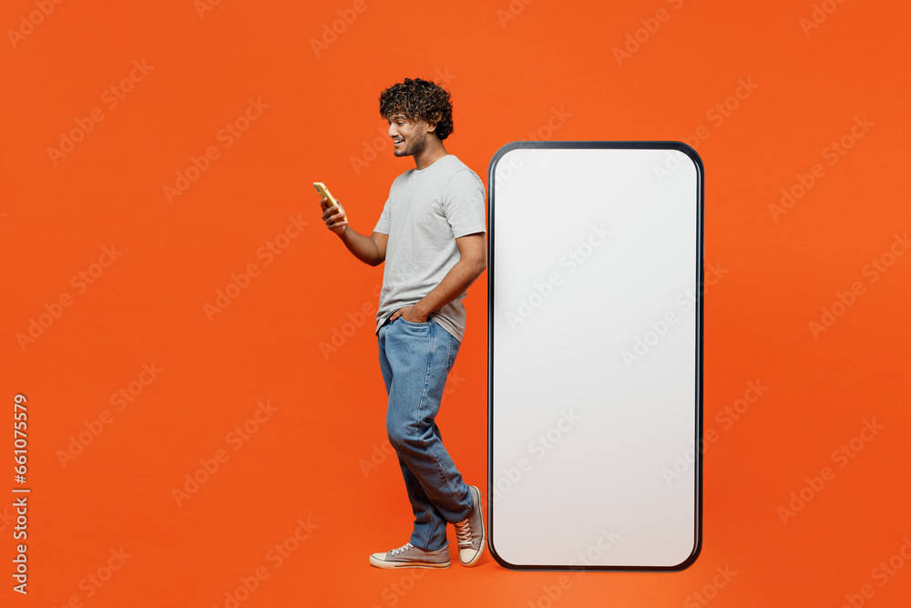 Full body side profile view young Indian man he wears t-shirt casual clothes big huge blank screen mobile cell phone with workspace mockup area use smartphone isolated on orange red color background. - obrazy, fototapety, plakaty 