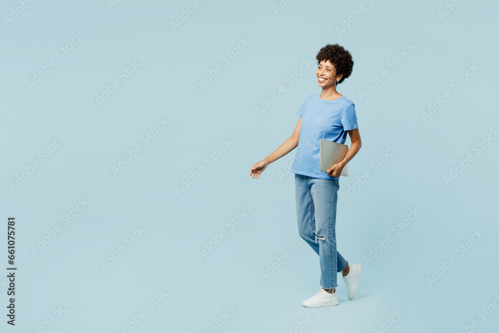 Full body side profile view smiling happy young IT woman of African American ethnicity wear t-shirt casual clothes hold closed laptop pc computer isolated on plain pastel light blue cyan background. Full body side profile view smiling happy young IT woman of African American ethnicity wear t-shirt casual clothes hold closed laptop pc computer isolated on plain pastel light blue cyan background.