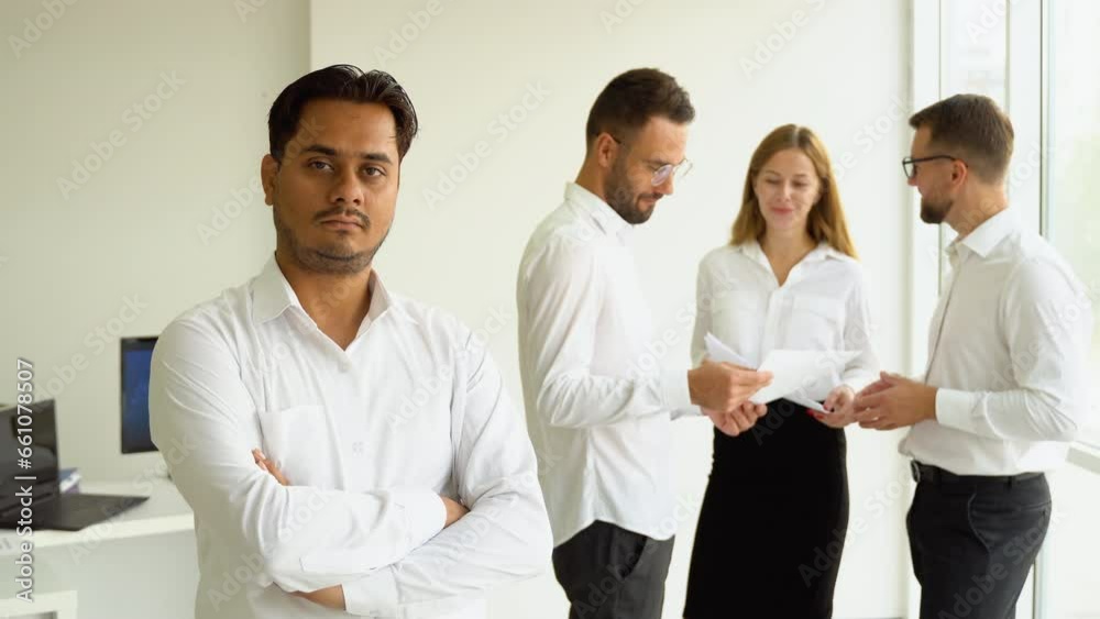 Confident mature Indian businessman with crossed arms looking at camera standing in office at team meeting Confident mature Indian businessman with crossed arms looking at camera standing in office at team meeting