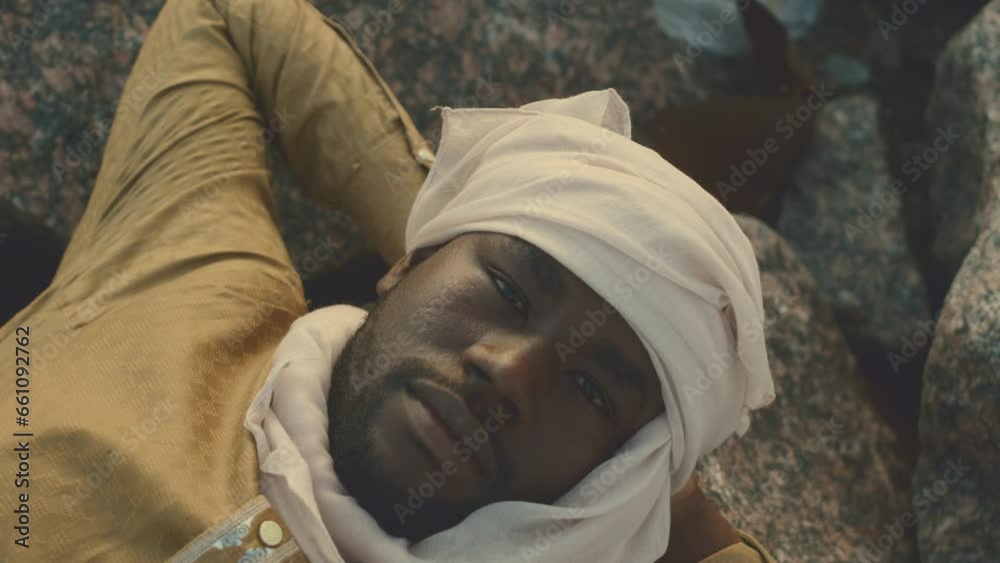 Zoom out portrait of handsome African American man wearing headscarf lying with his hands behind his head on beach stones and looking at camera Zoom out portrait of handsome African American man wearing headscarf lying with his hands behind his head on beach stones and looking at camera