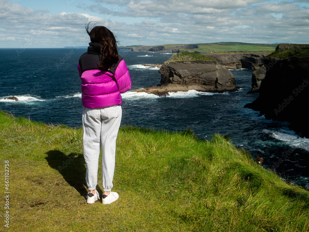 Girl looking at stunning nature scene with cliffs, Atlantic ocean and low cloudy sky on sunny day. Kilkee area, Ireland. Travel, tourism and sightseeing concept. Irish landscape and coastline.