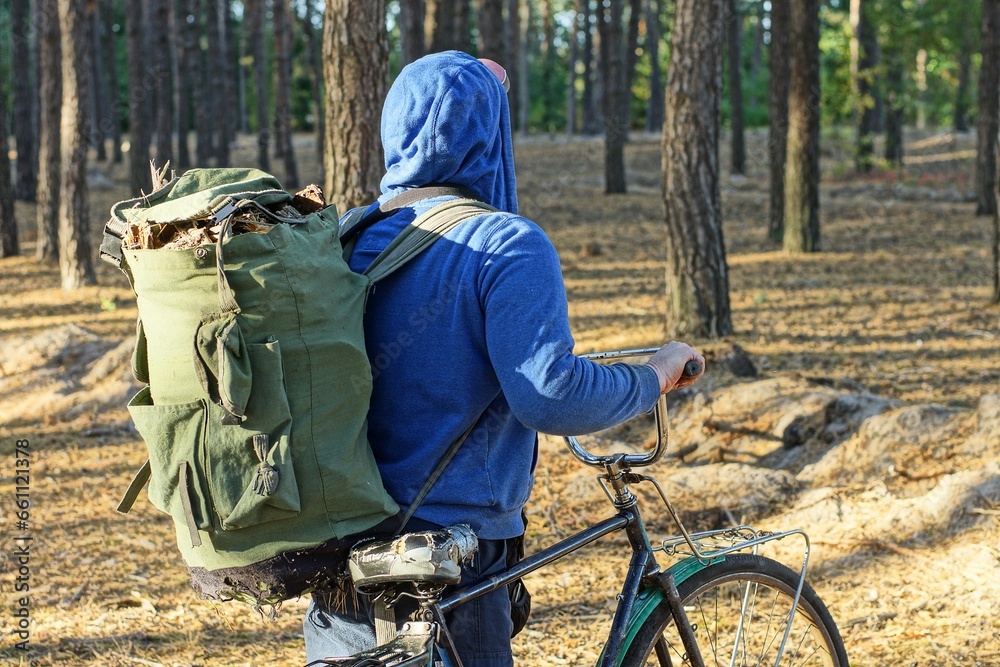 one man in a blue stylish raglan and hood with a large green full backpack with wooden sticks for a fire stands on the ground in the forest among the trees with an old iron black retro classic bicycle