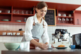 Cheerful female barista with cup of coffee on counter, preparing order for customer, happy service works in cafe, young small business startup entrepreneur
