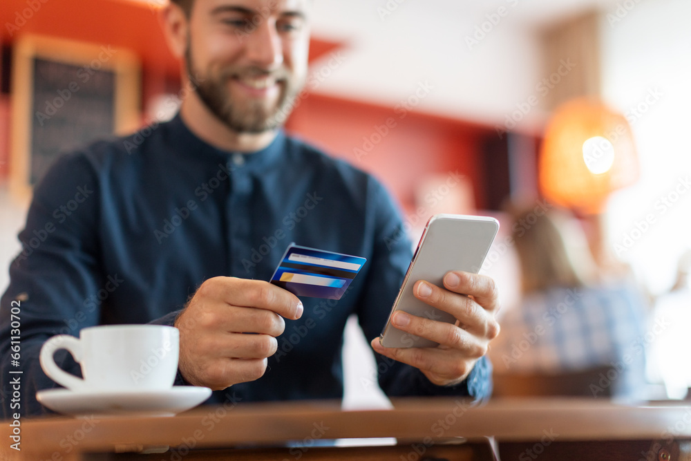 Handsome smiling european man with credit card and smartphone at cafe, drinking coffee and shopping online, purchasing in internet, focus on hands Handsome smiling european man with credit card and smartphone at cafe, drinking coffee and shopping online, purchasing in internet, focus on hands