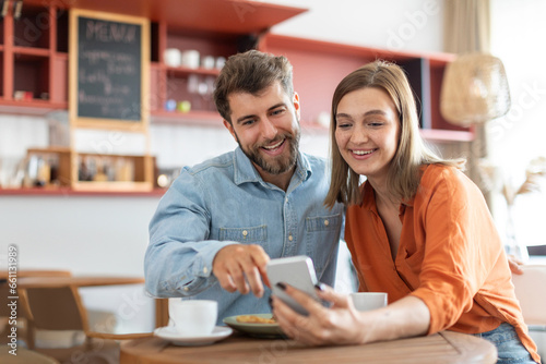 Wallpaper Mural Happy european couple using smartphone, sitting in cafe together on daytime, surfing internet or shopping online. Modern relationships concept Torontodigital.ca