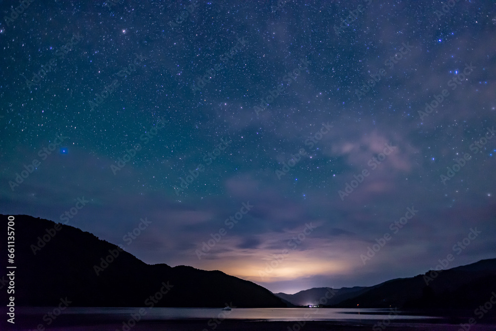 starry sky above the shape of mountains with the light of a city in a fjord in the Marlborough Sounds, New Zealand	