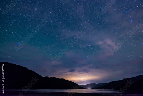 starry sky above the shape of mountains with the light of a city in a fjord in the Marlborough Sounds, New Zealand 