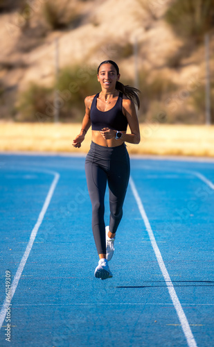 Beautiful slim young tanned runner girl  dressed in tight sportswear  smiling happily and enjoying running energetically on a blue track  approaching and looking at the camera.
