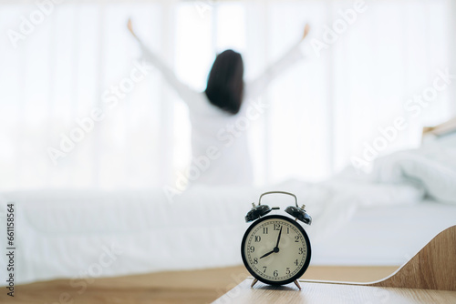 Close up of alarm clock on bedside table. Young Woman Waking Up with Happiness in Cozy Bedroom at Home. Beautiful Girl Smiling and Relaxing in Bed after Waking Up.