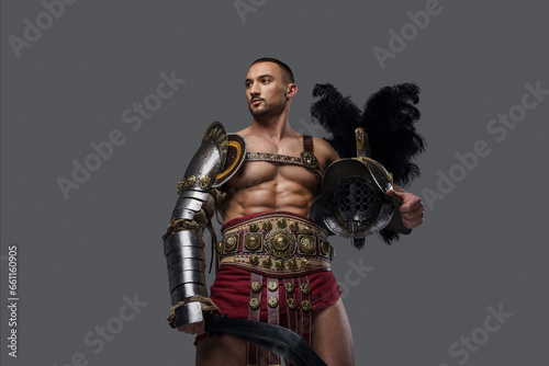 Muscular gladiator in graceful lightweight armor strikes a pose while holding a gladius and helmet, on a grey background