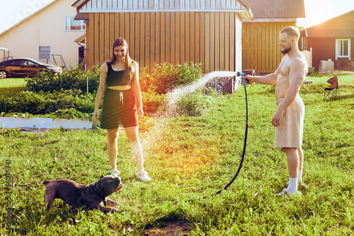 Pet owner waters dog with garden hose, which amuses animal and young woman of about 20 years old photo