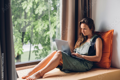 Young woman uses a laptop computer while sitting on a windowsill in hotel