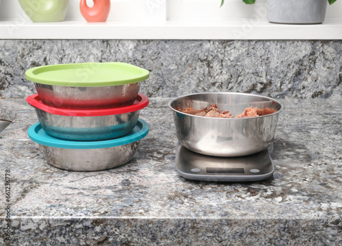 Raw food meal prep for dog in kitchen. Multiple pet food dishes and with scale. Measured ground chicken meat in portion and storing in stainless steel bowls with lids. Selective focus.