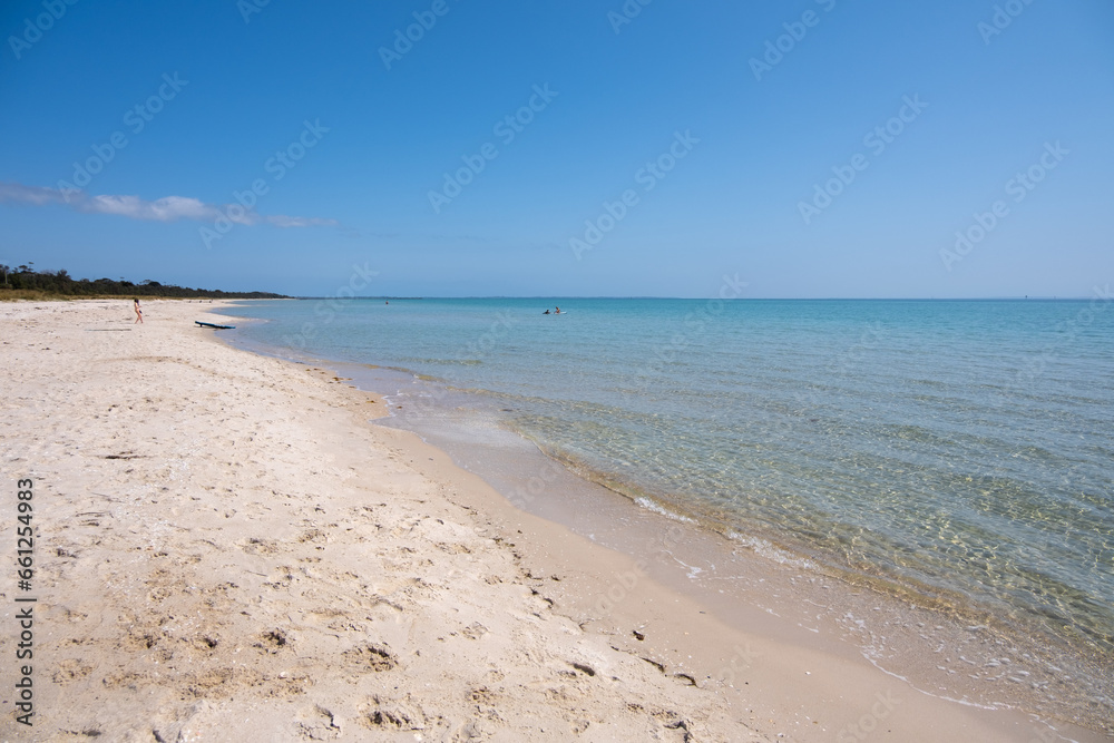 Background texture of clean white sands and shallow crystal clear shallow water against blue sky with a few unidentified people playing in the distance. Rosebud Beach, Mornington Peninsula, Australia