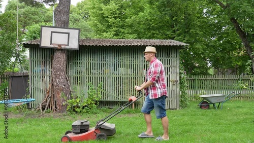 Father in a checkered shirt and a straw hat mows the lawn while walking up and down his little garden, with basketball hoop and a swing hung in a pine tree.