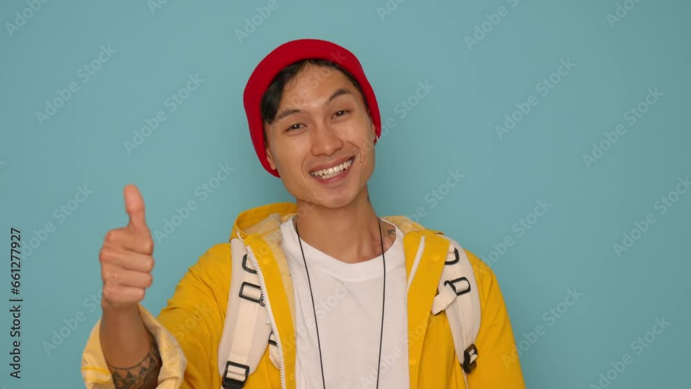 young attractive and happy Asian Korean man in red beanie and yellow hoodie smiling cheerful giving thumbs up success gesture isolated over blue background young attractive and happy Asian Korean man in red beanie and yellow hoodie smiling cheerful giving thumbs up success gesture isolated over blue background