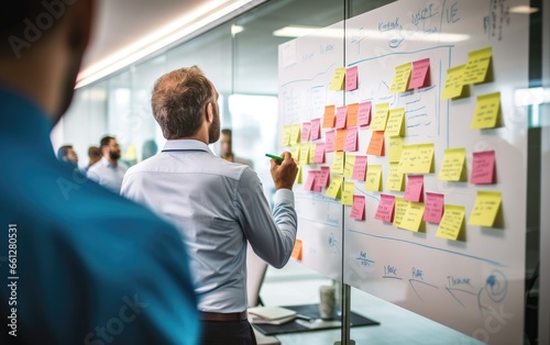 An individual stands before a whiteboard, brainstorming solutions to a challenge. Around them, colleagues engage in discussions, offering suggestions photo