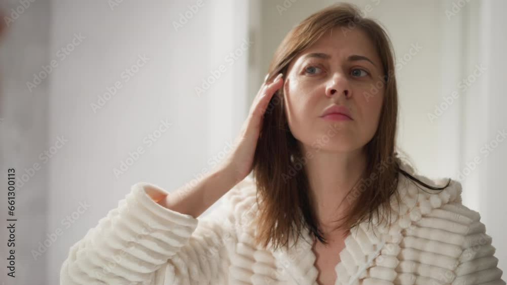 Woman in robe looks at face in mirror after going to spa center. Attractive lady with dark hair looks at effect after facial mask in spacious spa area Woman in robe looks at face in mirror after going to spa center. Attractive lady with dark hair looks at effect after facial mask in spacious spa area