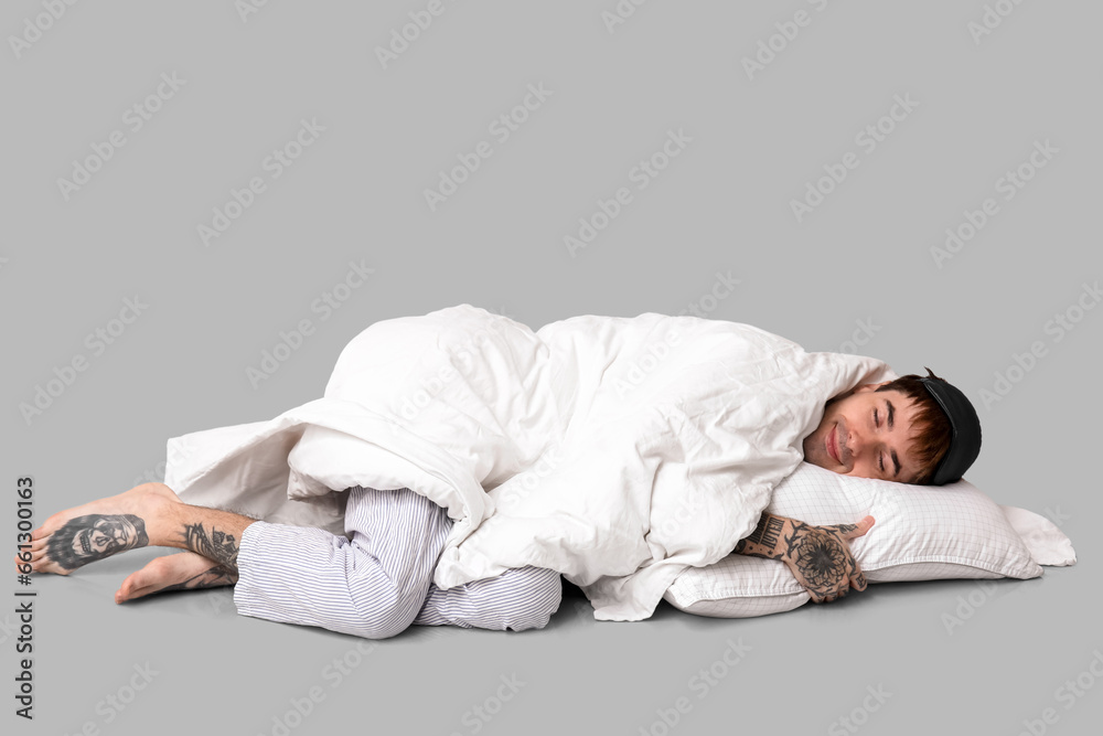 Tattooed young man in pajamas with bedding sleeping on grey background Tattooed young man in pajamas with bedding sleeping on grey background