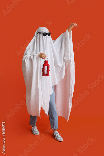 Young woman in Halloween costume of ghost and sunglasses holding lantern on color background