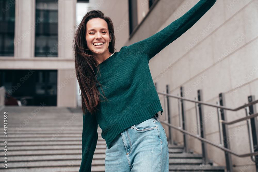 Fototapeta premium Street fashion portrait of beautiful caucasian woman walking outdoors. Beautiful young model in warm sweater. Fashion stylish pretty female posing in the street, having fun in city. Raising hands