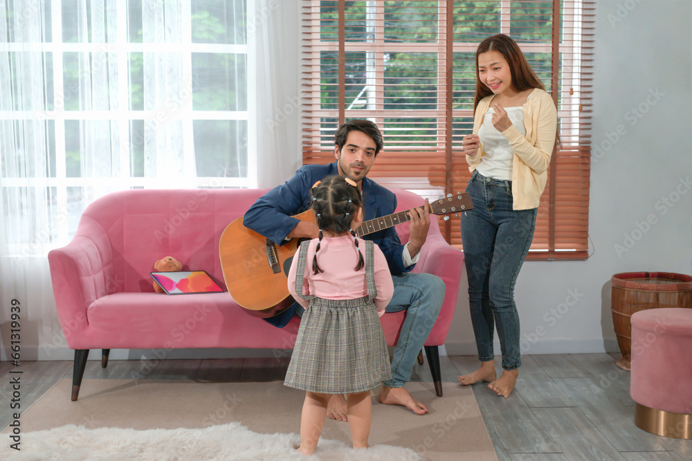 father sitting on sofa playing guitar,a little daughter looking at him while mother standing,sing a song,family spend time together in the living room,concept family,relationship,the love of a family father sitting on sofa playing guitar,a little daughter looking at him while mother standing,sing a song,family spend time together in the living room,concept family,relationship,the love of a family