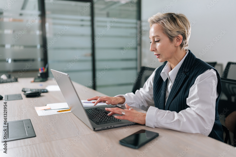 Side view of serious middle-aged business woman using typing on laptop computer sitting at workplace desk. Focused senior older employee 50s businesswoman executive working at home office. Side view of serious middle-aged business woman using typing on laptop computer sitting at workplace desk. Focused senior older employee 50s businesswoman executive working at home office.