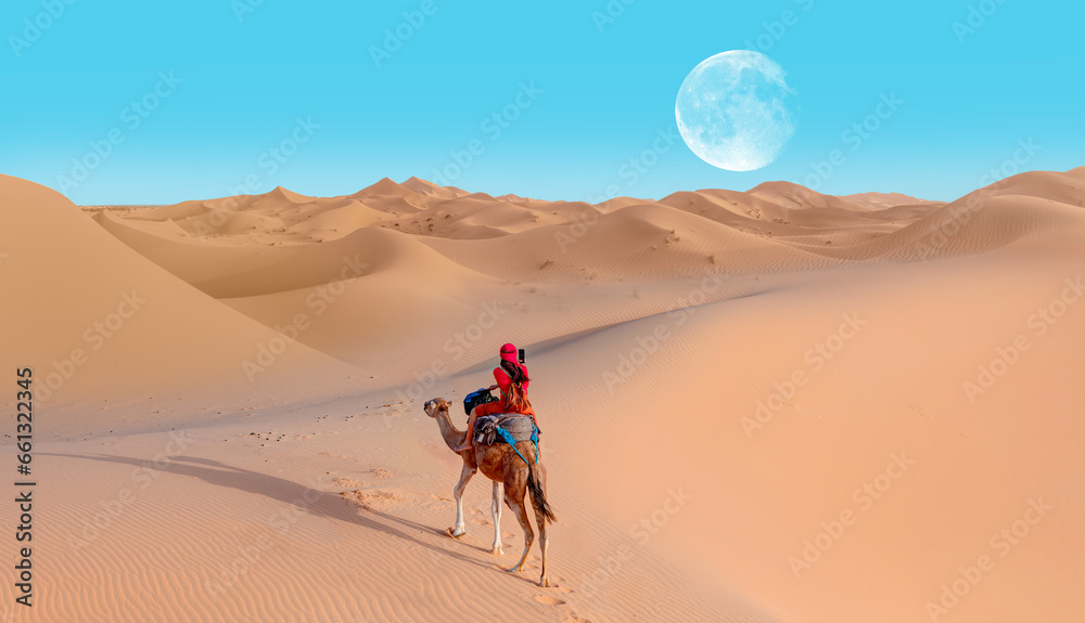 A woman in a red turban riding a camel across the thin sand dunes of the in Western Sahara Desert, Morocco, Africa A woman in a red turban riding a camel across the thin sand dunes of the in Western Sahara Desert, Morocco, Africa
