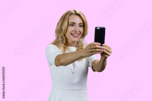 Portrait of happy blonde woman taking selfie on smartphone frontal camera. Isolated on pink.