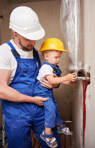 Male plumber holding little boy while child installing plumbing pipes in apartment under renovation. Father and son in safety helmet working on installation of sanitary equipment at home.