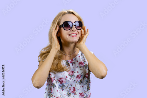 Happy cheerful mature blonde woman puts on sunglasses. Isolated on purple.