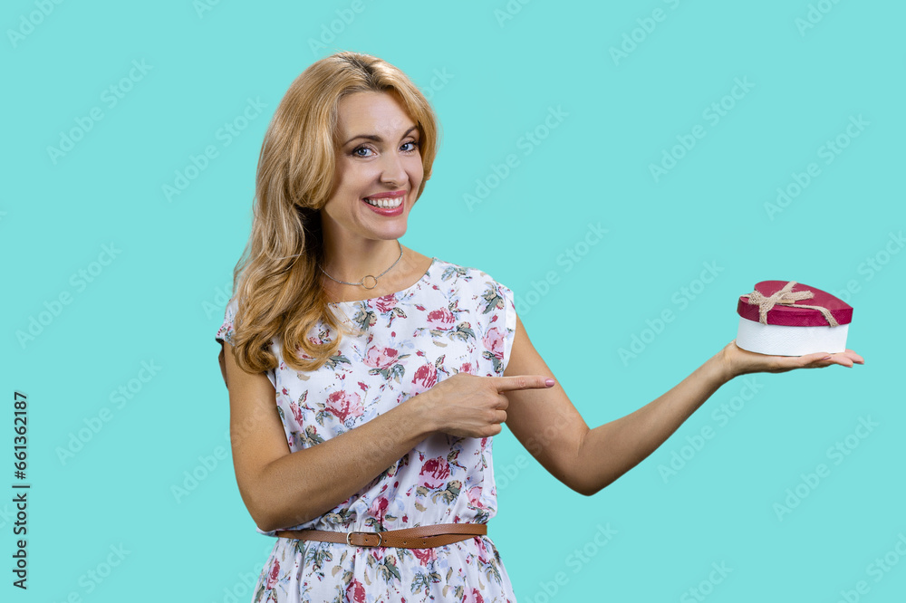 Blonde mature woman pointing at heart shape gift box. Isolated on turquoise. Blonde mature woman pointing at heart shape gift box. Isolated on turquoise.