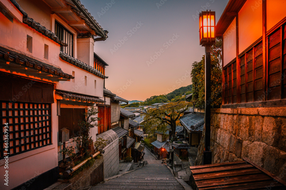 Fototapeta premium Kyoto City historic old town at sunrise with the warm blue sky in Japan, an empty downhill street with rustic wooden houses, cherry trees, and illuminated street light lamp