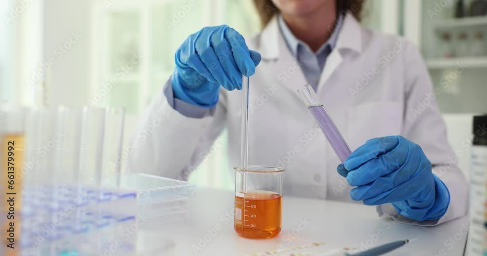 Chemist scientist holds and stirs flask with an orange liquid in hands in chemical laboratory. Laboratory assistant holding test tube with purple liquid Chemist scientist holds and stirs flask with an orange liquid in hands in chemical laboratory. Laboratory assistant holding test tube with purple liquid