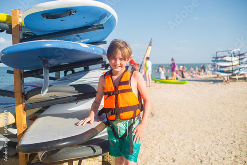 smiling child in a t-shirt with a backpack at the surf station on the beach in summer. joyful 10 year old child stands next to the windsurf boards © Ruslan Russland