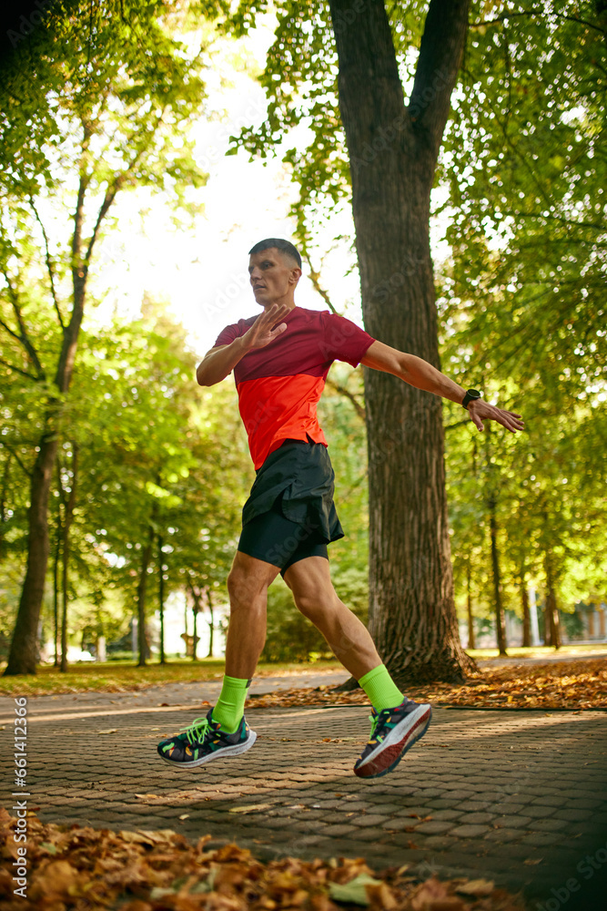 Muscular young man in sportswear doing warming-up muscles exercises, running, jumping on warm summer day in park. Concept of sport, active and healthy lifestyle, competition, dynamics, marathon
