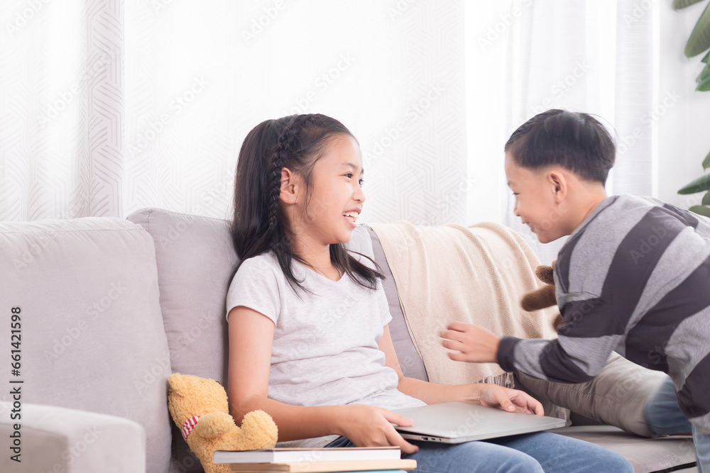 Two Asian siblings boy and girls having fun playing together on holiday at home while sister watches video games on modern technology laptop or doing homework on sofa, family relationship childhood