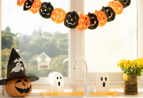 Halloween concept. A pumpkin with a painted face, a white ghost and a bouquet of yellow chrysanthemum flowers in a black vase against the background of a window in home interior.