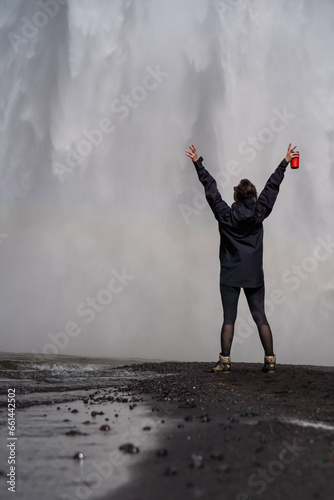 Traveller in black waterproof jacket near big waterfall with rainbow. Tourist girl holding red thermos cup with hot tea in hands. Travel in Iceland. Travel concept.