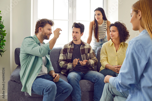 Group of friends gather at home and spend time together. Several people sitting on the couch and talking. Bunch of students playing charades. Young man tells a story or explains a word to his pals