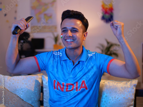 A young Indian man wearing a cricket jersey - watching a cricket match on TV  young cricket fan. An Indian man watching a live cricket match at home - wearing a jersey to support their team India  ... photo