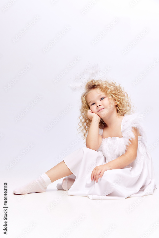 Cute, adorable little girl, child in white dress and angels halo sitting with dreamy face isolated on white studio background. Concept of childhood, imagination, fantasy, fashion and beauty, holidays Cute, adorable little girl, child in white dress and angels halo sitting with dreamy face isolated on white studio background. Concept of childhood, imagination, fantasy, fashion and beauty, holidays