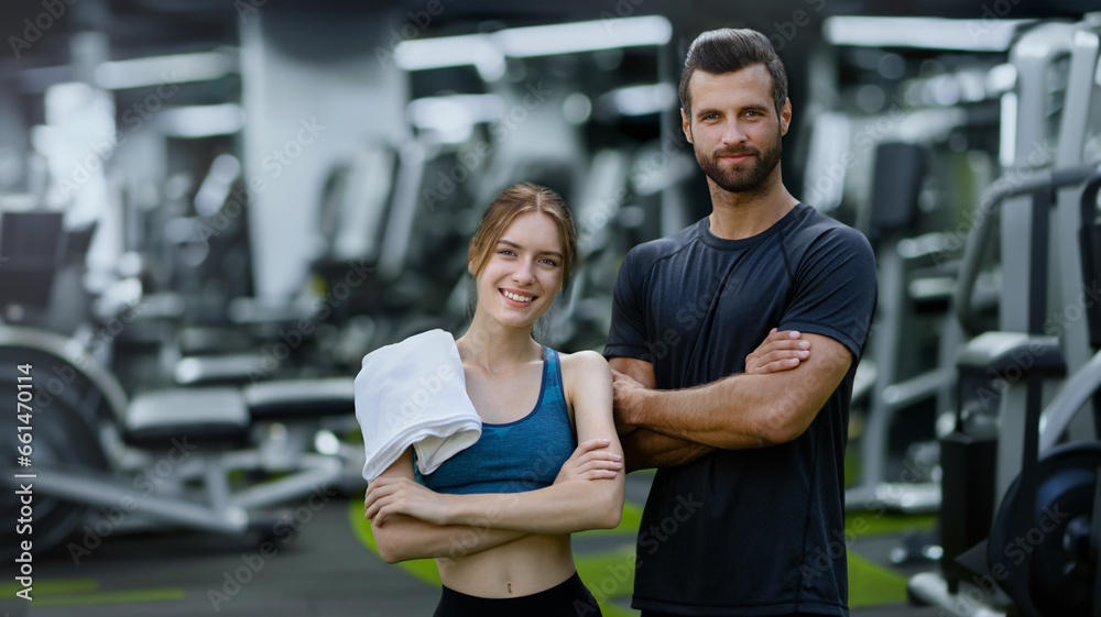 Fitness center training. Portrait image of smiling young couple, woman with man or male bearded coach trainer, standing with folded hands in sport machines gym. Fitness center training. Portrait image of smiling young couple, woman with man or male bearded coach trainer, standing with folded hands in sport machines gym.