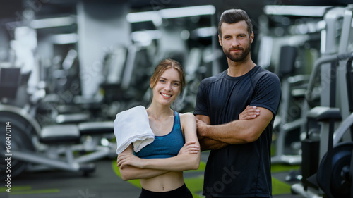 Fitness center training. Portrait image of smiling young couple, woman with man or male bearded coach trainer, standing with folded hands in sport machines gym.