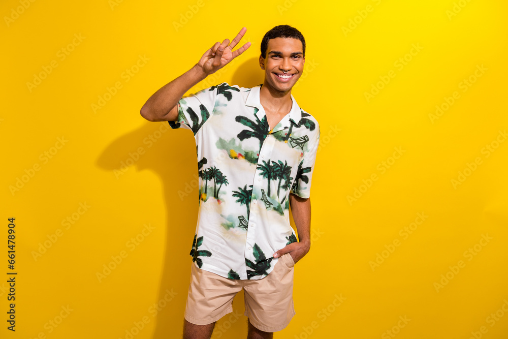 Portrait of pleasant funky guy with ring in nose wear print clothes showing v-sign hand in pocket isolated on yellow color background Portrait of pleasant funky guy with ring in nose wear print clothes showing v-sign hand in pocket isolated on yellow color background