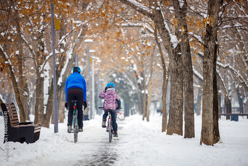 Father and child ride bicycles in winter. A man and his daughter ride bicycles along a bike path in a winter park. Back view