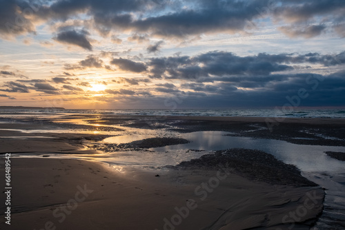A breathtaking moment captured as the golden sun sets over a picturesque French beach, casting warm hues on the shoreline, with small waves gently rolling in under dramatic clouds. Golden Sunset at